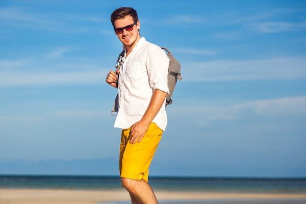 Outdoor fashion portrait of handsome man posing at amazing tropical beach, in nice sunny day, beautiful view on blue sky and ocean, wearing casual yellow sorts classic white shirt and sunglasses.